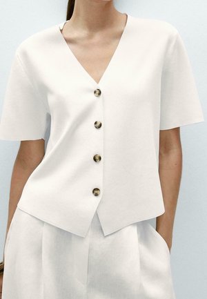 Femme portant un chemisier blanc à manches courtes avec un col en V et quatre boutons marron, et un pantalon blanc taille haute, les mains dans les poches.