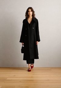 Schwarzer doppelt geknöpfter Mantel mit breiten Revers, langen Ärmeln und gerader Silhouette; kombiniert mit schwarzen Hosen und roten Schuhen.