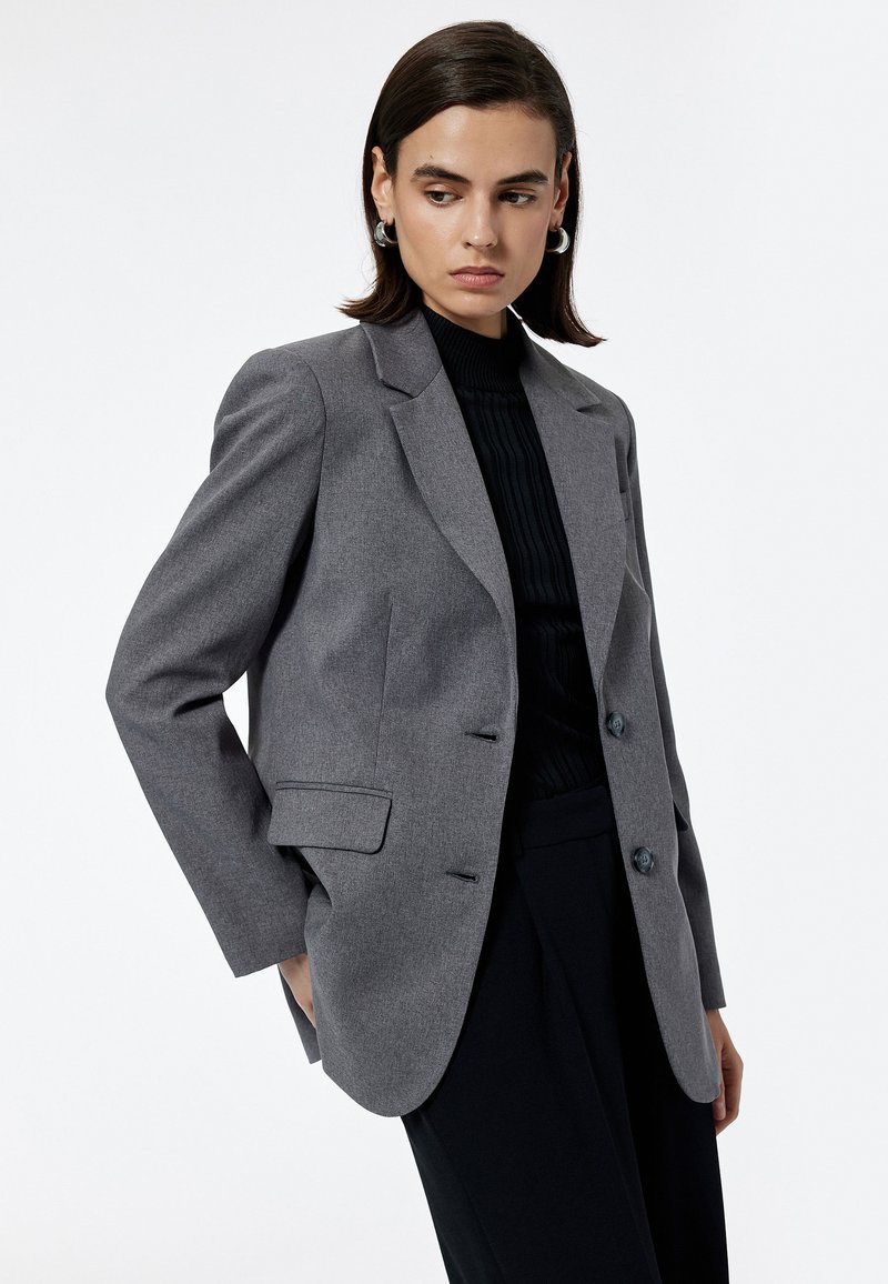 Kvinde med kort mørkt hår iført grå blazer over sort ribbet bluse og sorte bukser, kigger til venstre mod hvid baggrund.