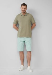 Khaki polo shirt met een eenvoudig ontwerp, gecombineerd met lichtgroene shorts en witte sneakers. Model staat met de handen op de rug geklapt.