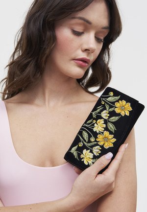 Žena u ružičastoj majici drži crni baršunasti pretinac sa žutim cvjetnim vezom, gleda prema dolje na pretinac.