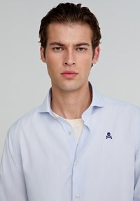 Hellblau gestreiftes Hemd mit Button-Down-Kragen, gesticktem Skull-Logo auf der Brust und glatter Baumwollstoffoberfläche.