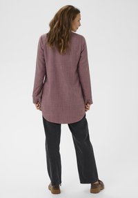 Ženska z dolgimi rjavimi lasmi, oblečena v dolge rukave mauve majice, črne široke hlače in rjave čevlje, stoji obrnjena stran od kamere proti beli ozadju.