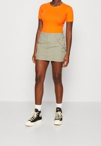 Khaki bavlněná sukna v cargo stylu se boční kapsou a řetízkovým detailem, spárovaná s přiléhavým jasně oranžovým topem a černými teniskami.