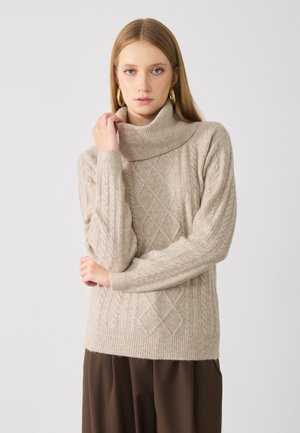 Maglione beige a collo alto in maglia con motivi a trecce e polsini a coste. La texture morbida enfatizza l'aspetto accogliente. Il modello posa con le braccia incrociate.