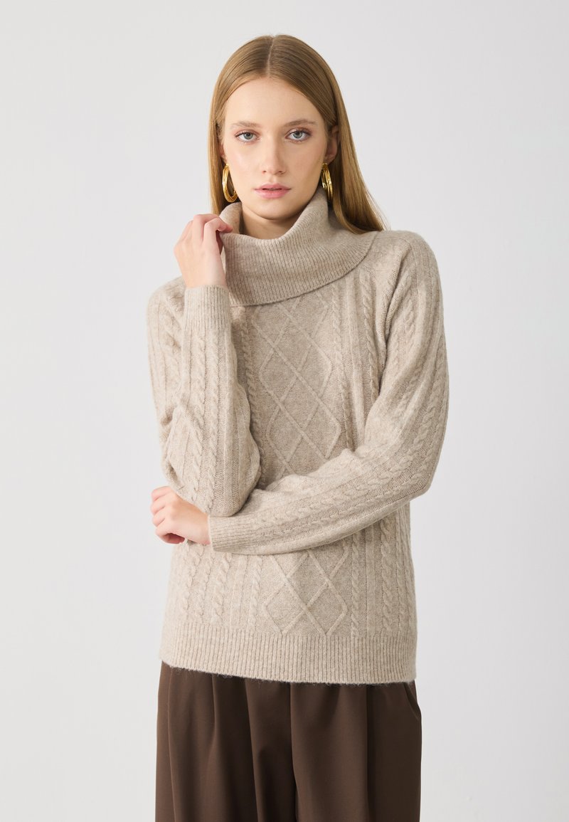 Beige adīta turtlenecks džemperis ar kabelīšu adījuma rakstiem un ribotām piedurknēm. Mīksta tekstūra uzlabo mājīguma izskatu. Modelis pozē ar rokām krusta.