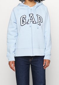 Light blue zip-up hoodie made of soft fabric, featuring black "GAP" lettering across the chest and a front kangaroo pocket.