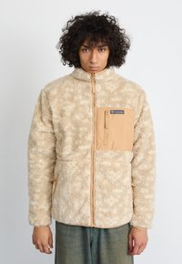 Person mit lockigem Haar trägt eine beige Fleecejacke mit Brusttasche und Reißverschluss und steht vor einem schlichten weißen Hintergrund.