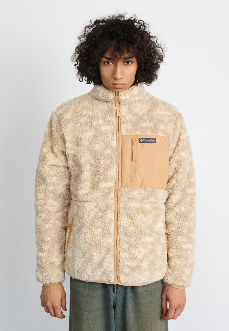 Person mit lockigem Haar trägt eine beige Fleecejacke mit Brusttasche und Reißverschluss und steht vor einem schlichten weißen Hintergrund.