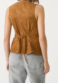 Bruine suède mouwloze top met een aangesloten lijfje en uitlopende zoom, voorzien van een strikdetail aan de achterkant en een gladde, zachte textuur.