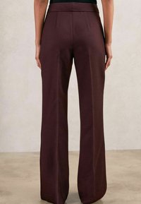 Magas derekú, bő szárú sötétbarna nadrágot viselő személy áll egy világos, egyszínű háttér előtt.