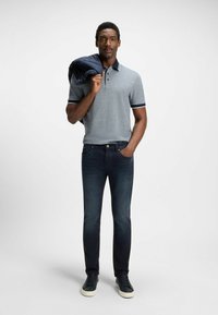 Camisa polo gris con acentos en navy, combinada con jeans azul oscuro y zapatillas negras. El modelo lleva una chaqueta navy sobre su hombro.