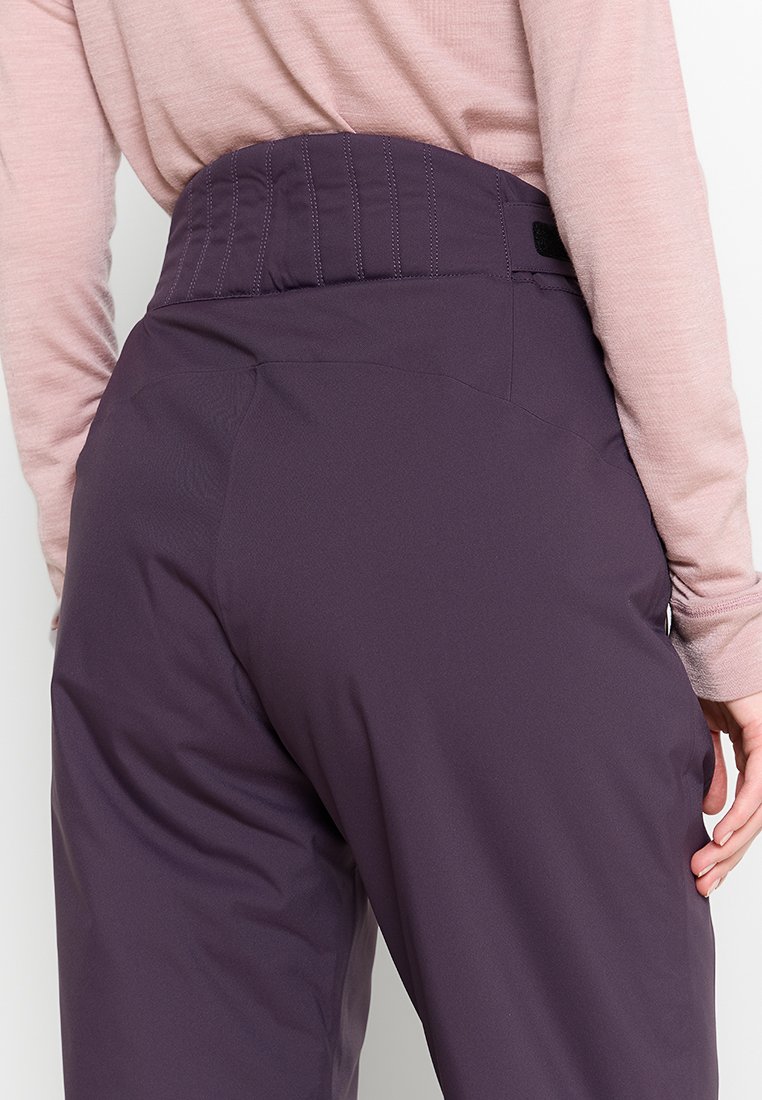 Frau trägt lila hochgeschnittene Hose mit gestepptem Bund und langärmligem, hellrosa Oberteil, Hand auf der Hüfte.