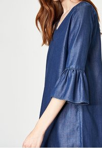 Džínové šaty v tmavě modré s výstřihem do V a krátkými rozšířenými rukávy. Mají lehký, hladký materiál a volný střih.