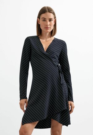 Femme portant une robe portefeuille noire à manches longues avec de fines rayures diagonales blanches et un ourlet asymétrique, debout sur un fond blanc uni.