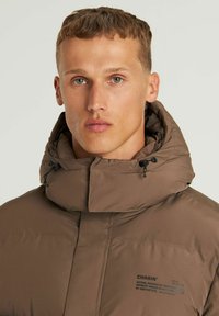 Chaqueta acolchada marrón con cuello alto, presenta un cordón, logotipo en el pecho y textura suave. Adecuada para usar en clima frío.