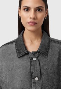 Giovane donna con capelli neri lisci che indossa piccoli orecchini a cerchio e una giacca di denim grigia con bottoni metallici, rivolta di fronte a uno sfondo semplice.
