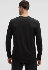 Muž otočený chrbtom, oblečený v jednoduchom čiernom tričku s dlhým rukávom a čiernych nohaviciach na neutrálnom pozadí.