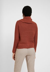 Pull tricoté couleur rouille avec un large col, des poignets et un ourlet côtelés, associé à un pantalon à motif check. Détail de tissu texturé visible.