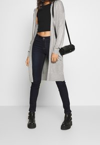 Kobieta ubrana w czarny krótki top, ciemne dopasowane dżinsy, szary długi kardigan, czarne botki za kostkę oraz czarną cylindryczną torebkę na ramię.