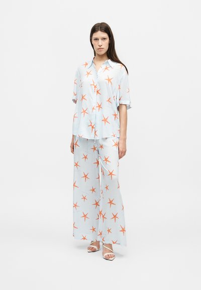 Femme debout portant une chemise ample assortie bleu clair et un pantalon à jambes larges avec un imprimé étoiles de mer orange et des sandales blanches à brides.