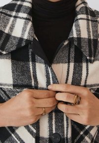Abrigo de lana a cuadros en blanco y negro con un gran cuello, cierre frontal de botones y un acabado texturizado. Anillos dorados en las manos completan el look.