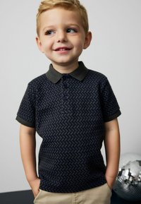 Polo di colore blu navy con un motivo geometrico, maniche corte e colletto verde scuro. Presenta tre bottoni e polsini arrotolati.