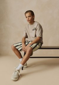 Beiges T-Shirt mit grünem und schwarzem Grafikdruck, kombiniert mit gestreiften Shorts in Pastellfarben. Hellgraue Sneakers mit cremefarbenen Akzenten.