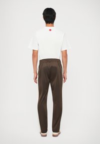 Witte t-shirt met korte mouwen en een rood rond logo op de achterkant, gecombineerd met bruine joggingbroek met witte zijstrepen.