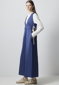 Rochie fără mâneci, de culoare albastră, cu decolteu în V adânc, talie elastică cu șnururi, buzunare laterale și o formă lejeră, fluidă.