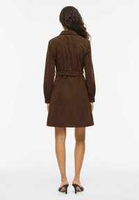 Pruun seemisnahast ümbermähitav kleit pikkade varrukatega, vöö ja kellukeseelikuga. Omaduseks on krae ja nööpidega mansetid. Sile tekstuur ja põlve keskmise pikkusega.