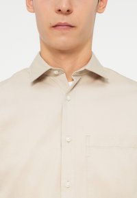 Μπεζ πουκάμισο με κουμπιά, με υφασμάτινη υφή, μυτερό γιακά και τσέπη στο αριστερό στήθος. Λευκά κουμπιά είναι ορατά κατά μήκος του κουμπώματος.