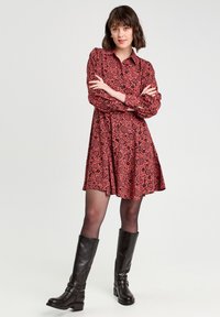 Robe à motifs floraux rouges avec un col, des manches longues, une taille cintrée et une jupe évasée, associée à des bottes noires jusqu'aux genoux.