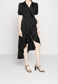 Robe portefeuille en satin noir avec manches ballon, ourlet asymétrique et détail noué à la taille. Associée à des sandales à talons hauts noires à brides.