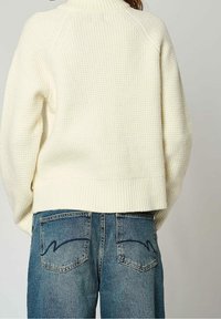 Pull tricot épais blanc avec un col montant, associé à un jean denim bleu avec des poches arrière incurvées et une texture délavée.