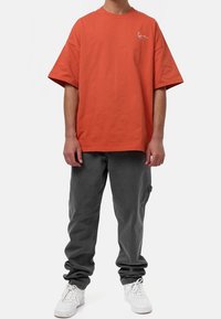 Oranssi puuvillateepaita, jossa on lyhyet hihat ja rento istuvuus; harmaat kapenevat housut, joissa on sivutaskut; valkoiset lenkkarit.