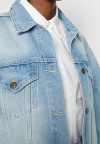 Light blue denim jacket with a classic collar, front pockets, and brass buttons. Features subtle stitching details and a slightly faded texture.