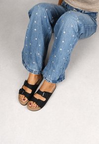 Schwarze Sandalen aus einem glatten Material, mit zwei verstellbaren Schnallen. Getragen mit blauen Jeans aus Denim, die mit silbernen Nieten verziert sind.
