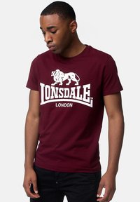 Tricou din bumbac burgundy cu un logo grafic alb reprezentând un leu, text curbat "LONSDALE LONDON" în partea de jos, și design cu guler rotund.
