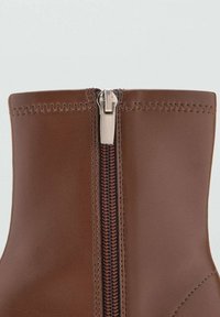 Botín de ante marrón con una cremallera plateada en la parte frontal, que presenta un hilo rojo en contraste a lo largo de la cremallera y una textura suave.