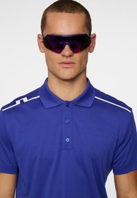 Blue polo shirt with three buttons and white shoulder stripes, paired with large, dark sunglasses featuring a purple tint and angular design.