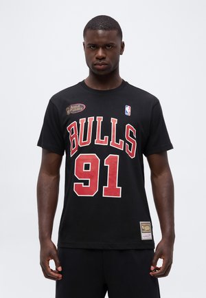 Νέος άνδρας που φοράει μαύρο μπλουζάκι Chicago Bulls NBA Finals με τον αριθμό 91, στέκεται μπροστά σε απλό ανοιχτόφωτο φόντο.