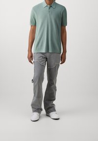 Polo à manches courtes vert clair avec un motif texturé, associé à un pantalon cargo gris avec des fermetures éclair latérales et des baskets blanches.