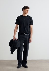 Νεαρός άνδρας όρθιος, φοράει μαύρο μπλουζάκι και τζιν, κρατώντας ένα μαύρο μπουφάν στο αριστερό του χέρι, κοιτάζει προς τα δεξιά του.