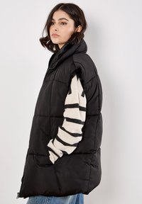 Zwarte puffer vest met hoge kraag, gewatteerde textuur en zijzakken, gedragen over een gestreept langmouwshirt met een relaxte pasvorm.