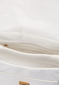 Vit konstläderhandväska med flera fack, ett blixtlåsområde och detaljer i gyllene färg. Slät, polerad yta.