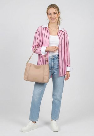 Jeune femme en chemise rayée rose, haut blanc, jean bleu clair, baskets blanches tenant un sac à main beige sur son bras, souriante sur fond gris.