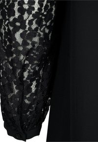 Manche en dentelle noire avec motif floral fixée à un tissu noir uni, manchette boutonnée visible au poignet.