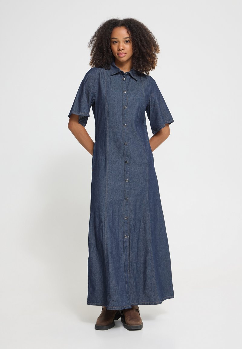 Femme debout, les mains dans les poches, portant une longue robe en jean à boutons et des bottes brunes, sur un fond blanc uni.