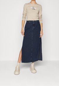 Beige långärmad skjorta med tryckt logotyp, kombinerad med en mörk denim maxi kjol med en sidoklyvning, och ljusa sneakers.